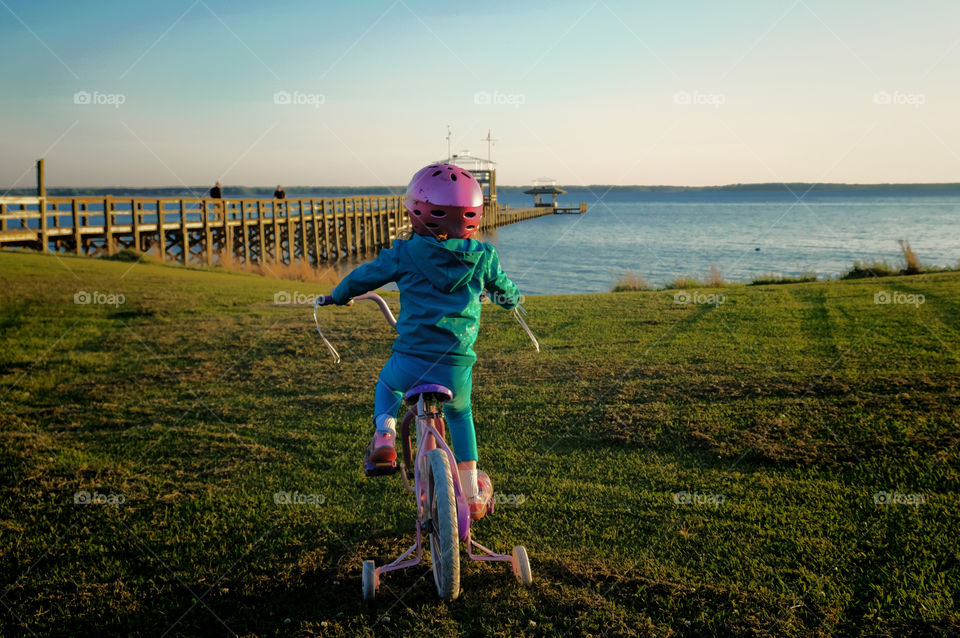Little girl riding her bicycle with training wheels towards the pier or dock over the Neuse River estuary in Arapahoe North Carolina. 