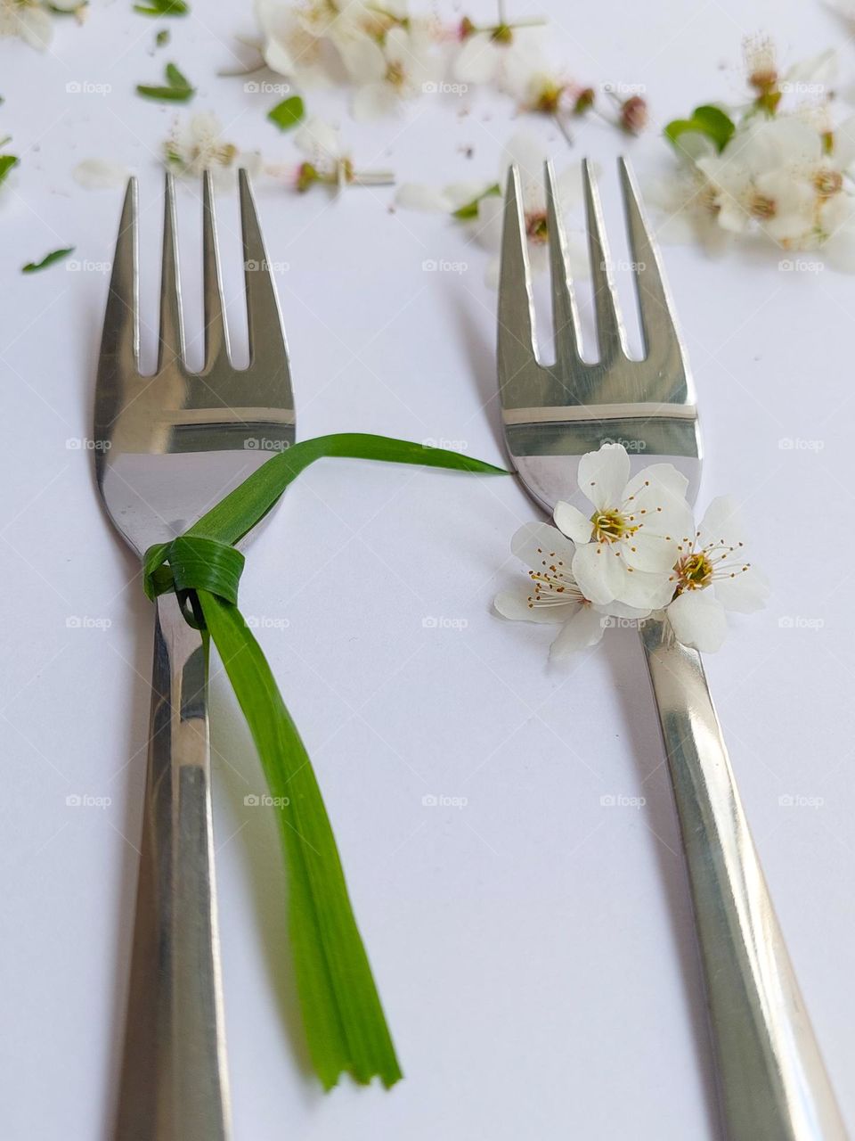 Two metal forks decorated with flowers and a grass tie,  as a woman and a man,  on a white table decorated with flowers