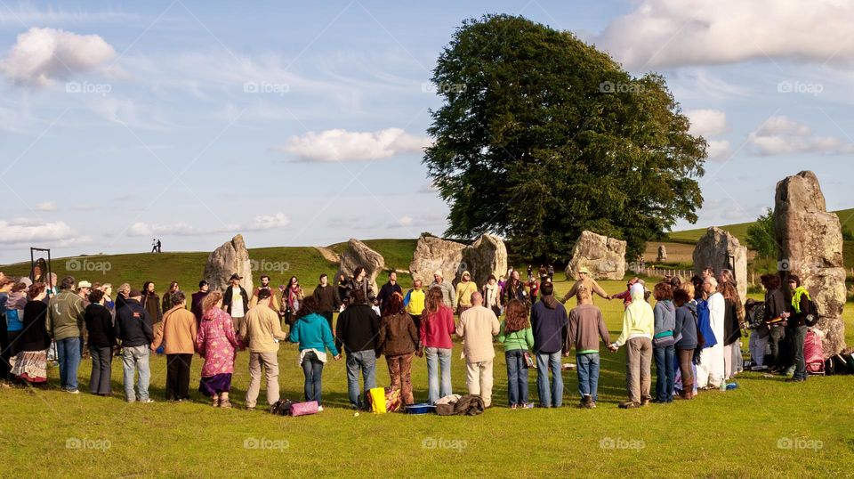 A crowd of people join hands around the standing stones at Avebury, UK