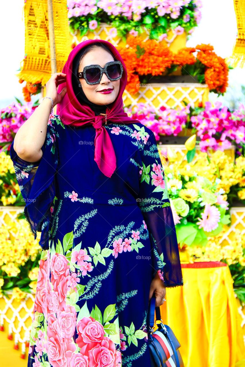 Portrait of a beautiful mother wearing a red hijab, wearing a floral patterned robe and wearing black glasses, taking a photo with a happy expression.