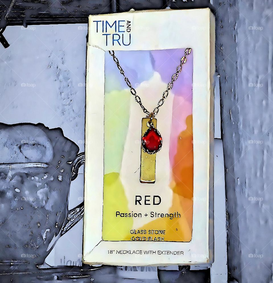 A beautiful sparkling red stone, dazzles reflecting on the gold rectangular plate of the necklace in a white box with a colorful mosaic background.