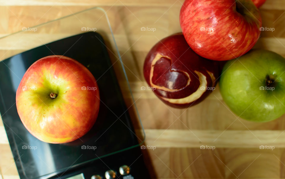 Apple on a scale on table next to different colored apples with heart shaped design carved into red apple for conceptual healthy choices and diet photography 