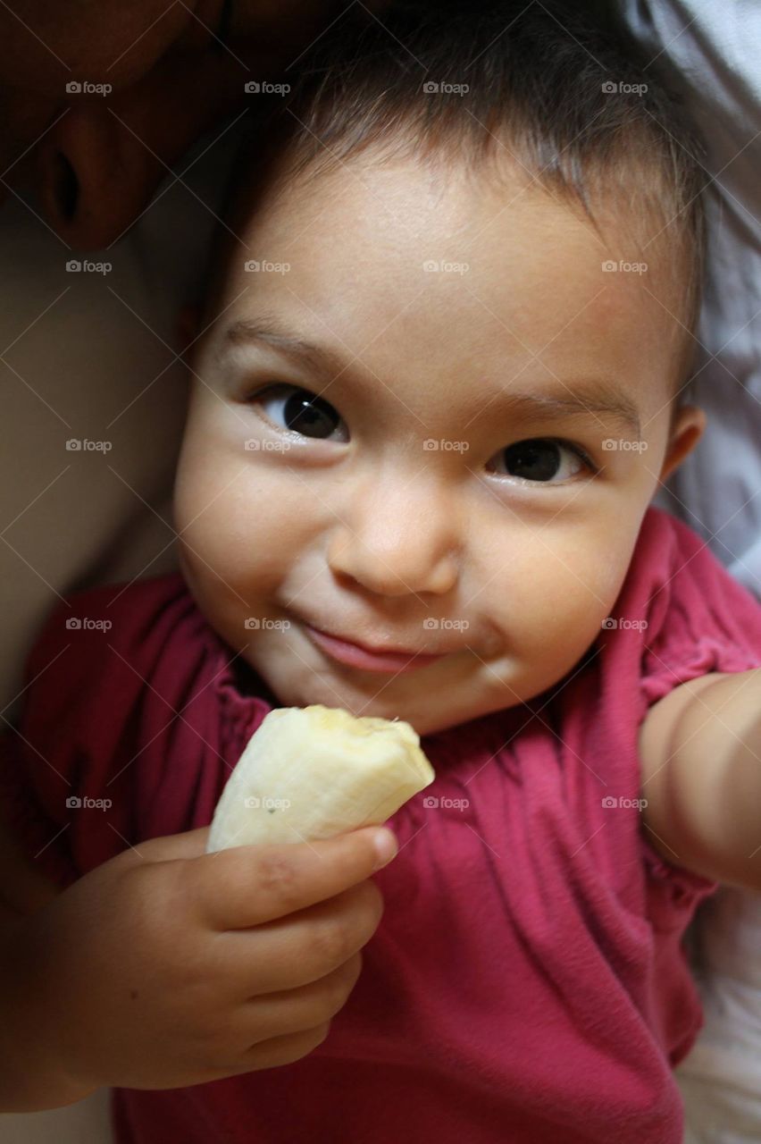 Cute toddler is eating banana in bed