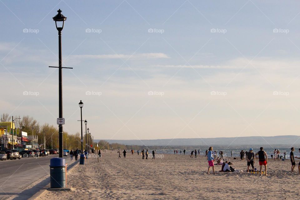 People on a brown sandy beach at sunset with blue sky and white clouds and a row of lampposts 
