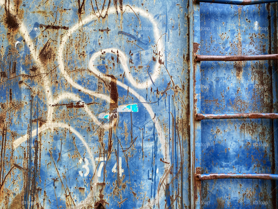Side of a rusty trash dumpster with graffiti and a ladder