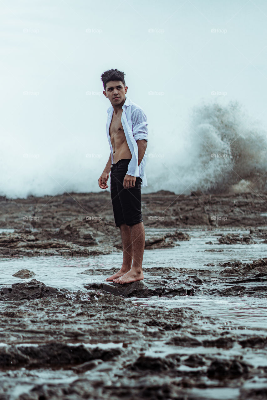 Boy on the beach with a wave breaking behind the rocks. Photography edited in soft colors