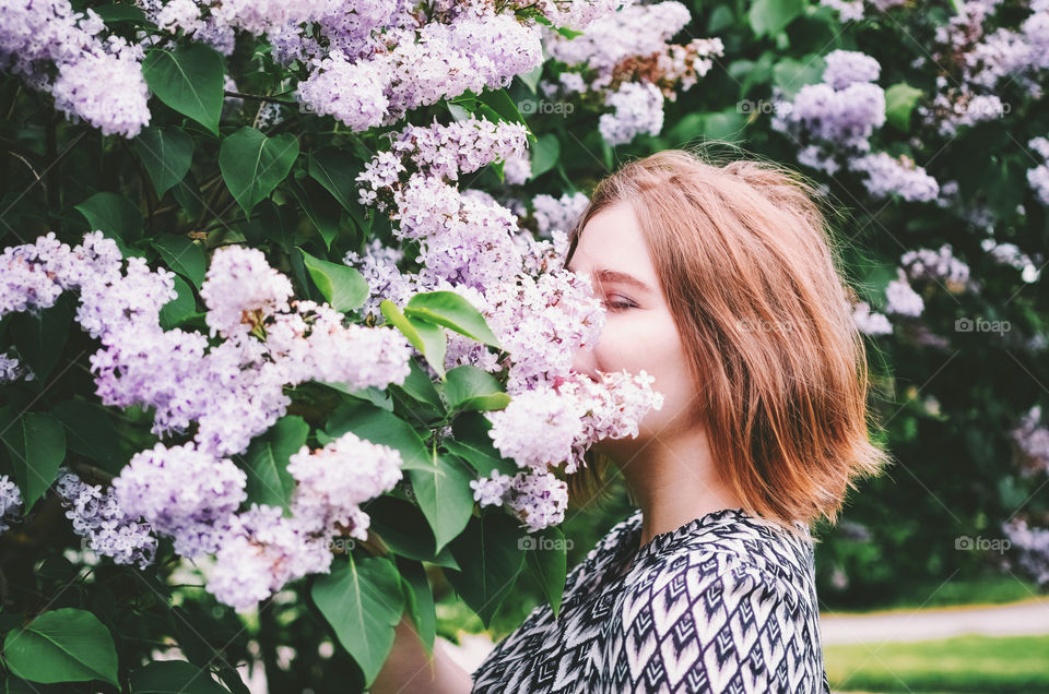 Portrait of a beautiful ginger girl sniffing blooming lilac flowers in a sunny summer park.