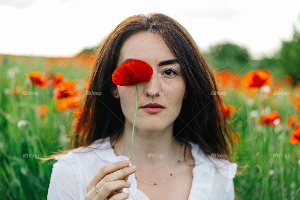 Beautiful woman holding a poppy flower in front of her eye, while being surrounded by a field of poppy flowers.