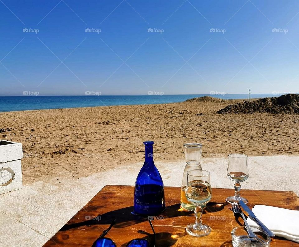 Love Romantic day by the sea. A table with a bottle of wine and two glasses of wine. In the background, warm sand, blue sky, which merges with the blue sky on the horizon.
