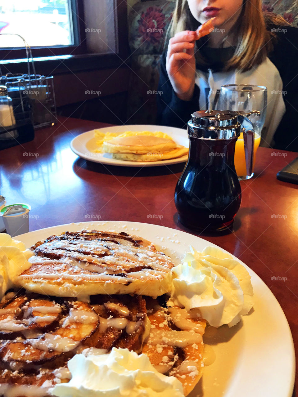 In the foreground; Cinnamon roll pancakes with sweet cream cheese sauce & whipped cream and across the table; pancakes, syrup and my daughter munching a piece of crispy bacon! We love  brunch!!! 🥞