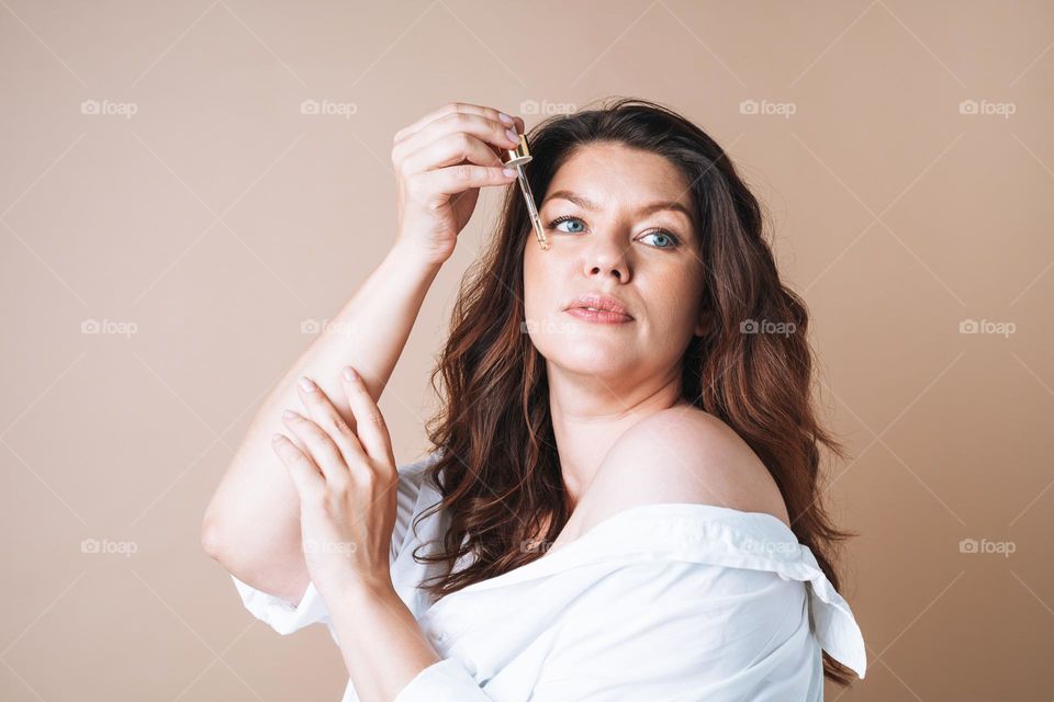 Beauty portrait of body positive woman with dark hair with pipette with organic oil on clean fresh skin face and hands on beige background