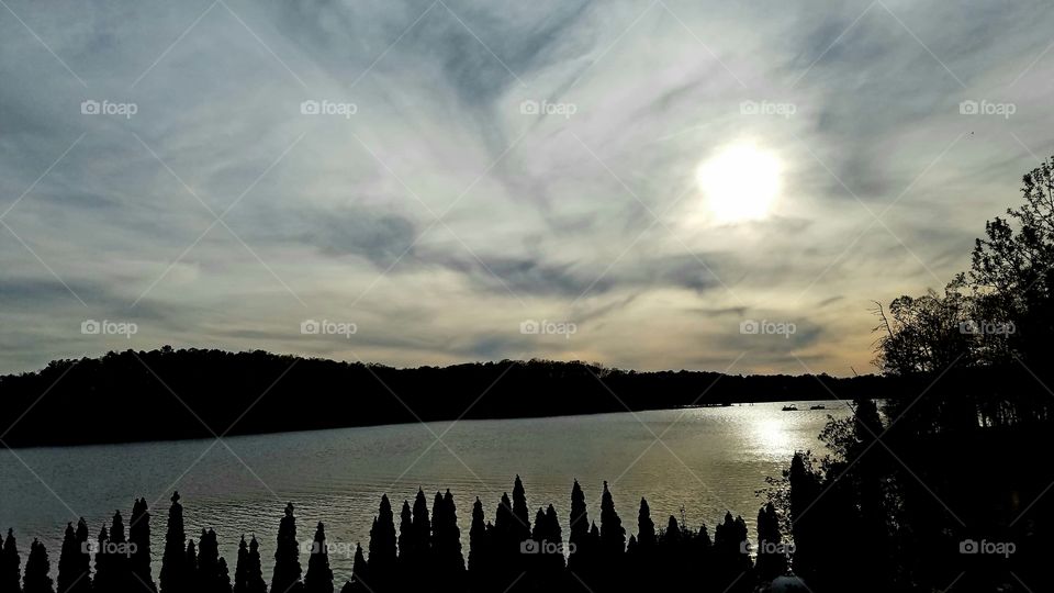 sun beginning its descent over lake surrounded by clouds.