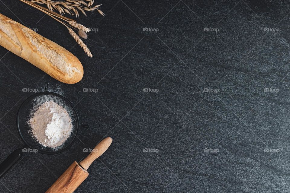 A French baguette, a rock, a sieve with flour and a bouquet of ears lie on the left on a black stone background with copy space on the right, a flat lay close-up. The concept of baking bread.