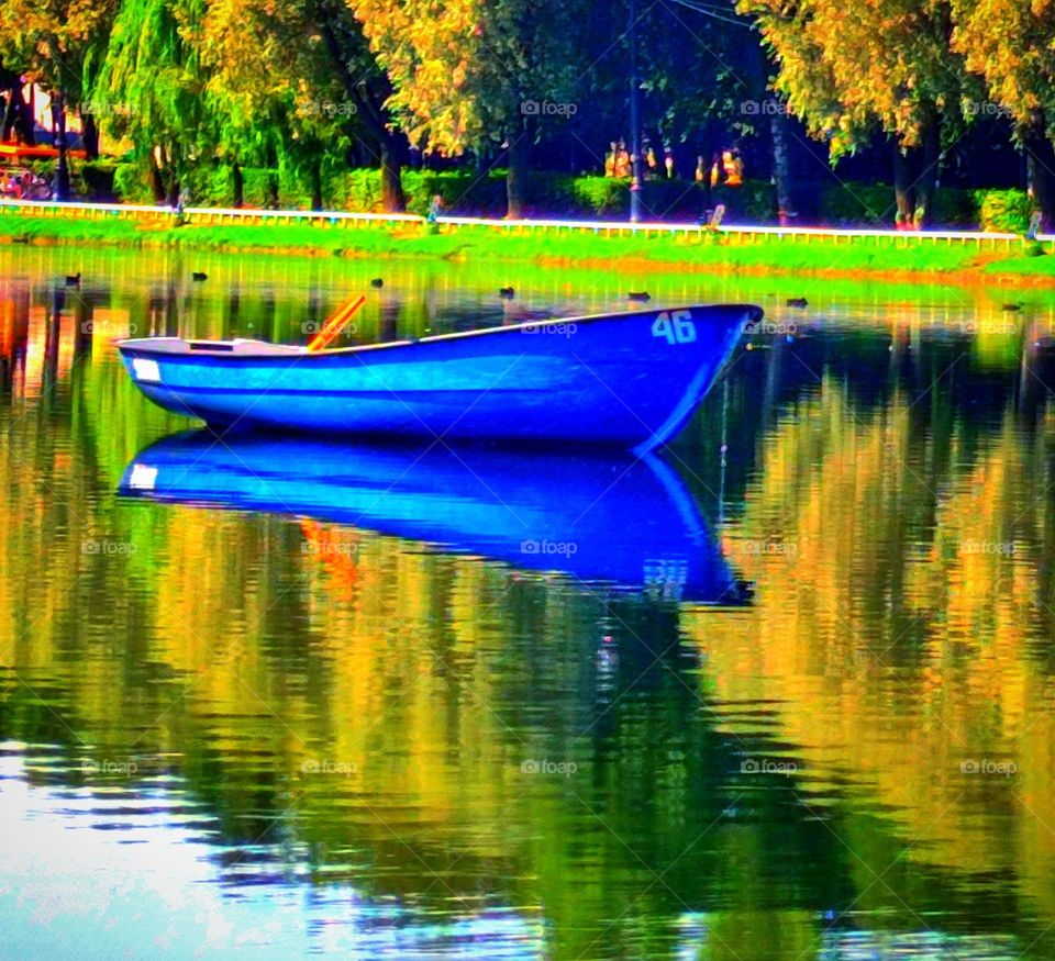 A boat in the middle of a pond with scales.  Autumn trees and a boat are reflected in the water