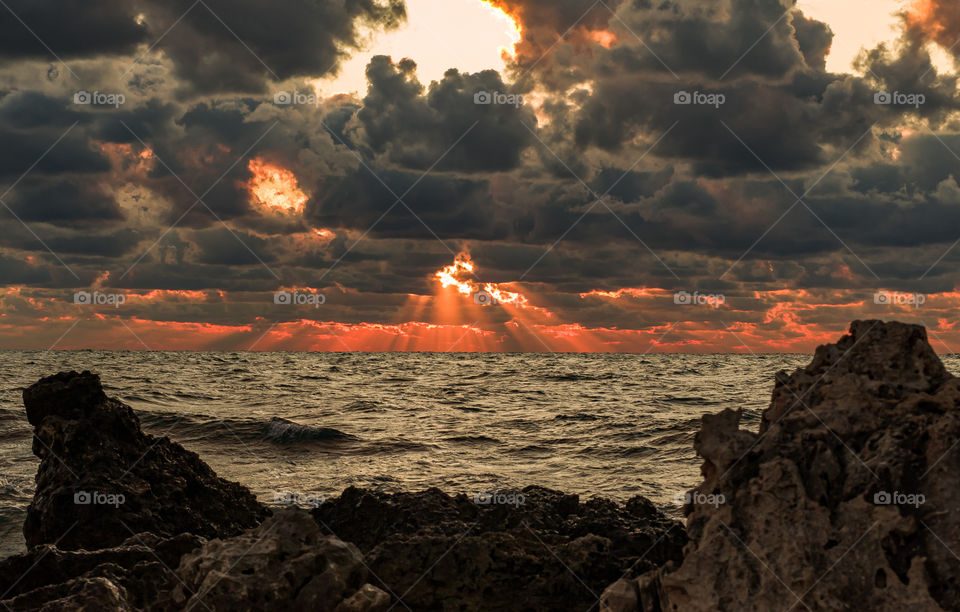 seascape with stones in the foreground and the rays of the setting sun breaking through the clouds in the background