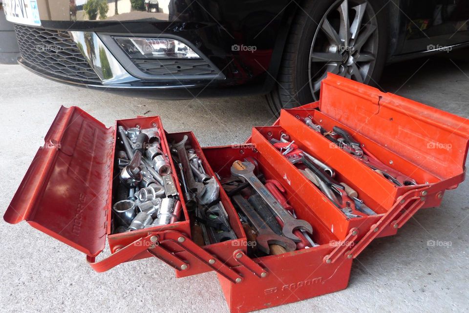 In a garage with a cement floor, a red tool box is open on the ground in front of a black car (at hood level). It is filled with various metal tools.