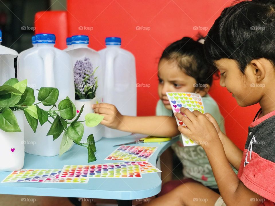 Two kids are doing craft with leftover plastic milk bottles, they making flower pots with plastic milk bottles and sticks some sticker’s on the bottles and they are enjoying and smiles by doing the craft.Kids must teach howto reuse the waste material