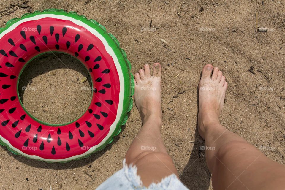 female legs and a swimming ring in the form of a watermelon on a sandy beach near the sea