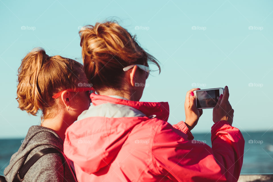 Back view of mother and her daughter taking a photos over sea during summer vacation using smartphone camera