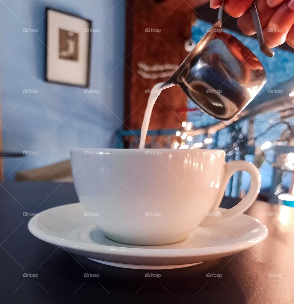 Cup of coffee with a hand spilling milk on top with a small silver milk jug, in a nice coffee shop with lights