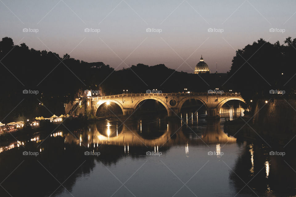 The ponte sisto bridge in Rome over the tiber river at night. it is nicely lit and in the distance there is a basilic visible. at the riverside there is a market.