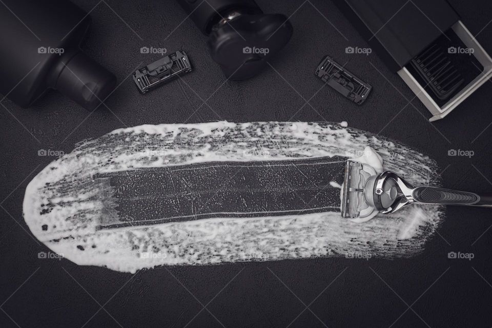 A swab of shaving foam with a razor, razor heads, a comb in a box, an electric razor and a bottle of aftershave cream on a black background with copy space in the center, flat lay close-up. The concept of a man barbershop, man shaving, beard care.