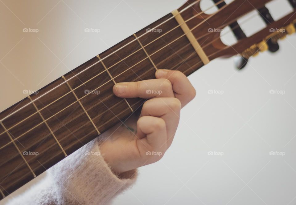 One hand of a little caucasian girl holds a guitar and picks the strings with her fingers while sitting on the sofa in the room in the morning and learns notes against the background of a yellow wall, close-up side view. Music education concept.