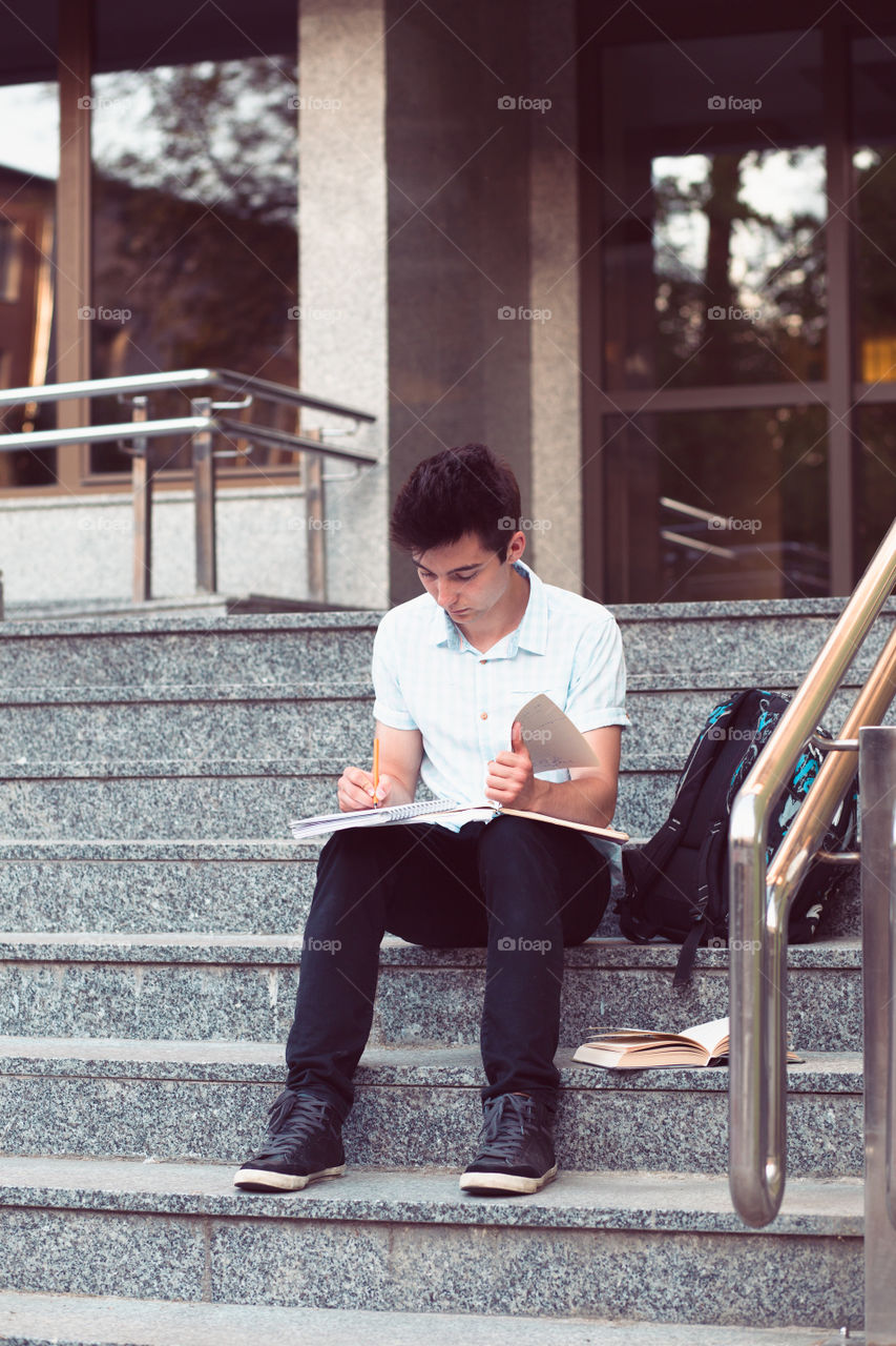 Student making a notes sitting on stairs outside of university. Young boy wearing blue shirt and dark jeans