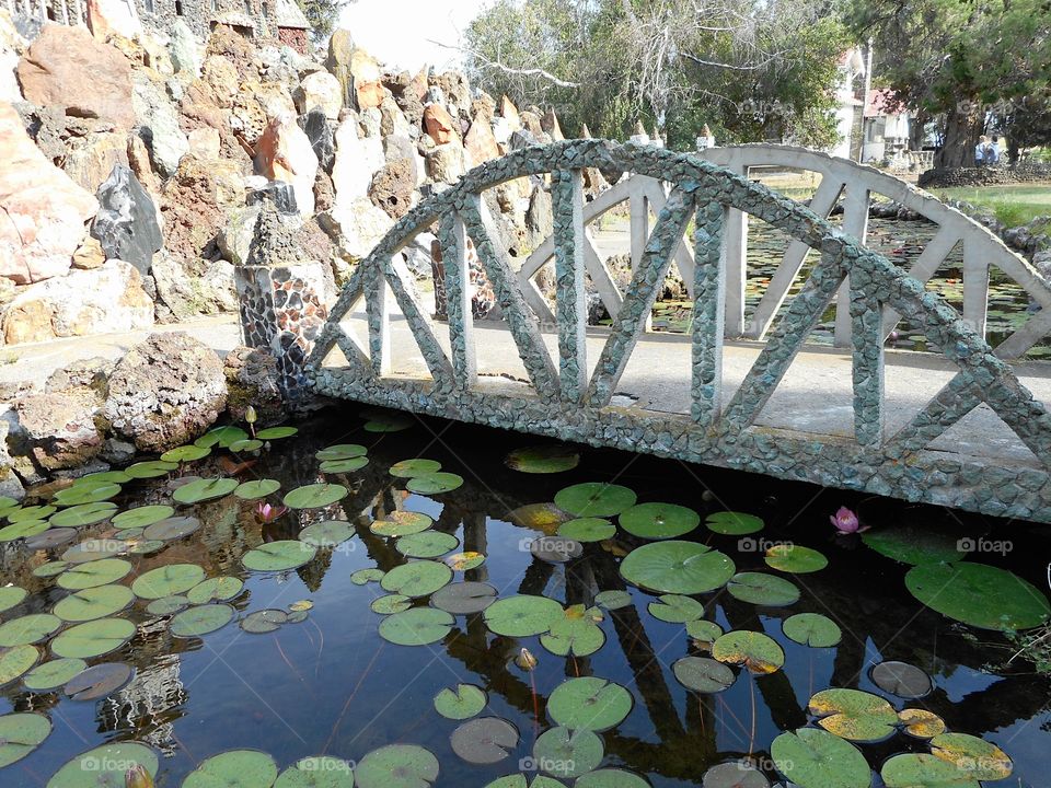 A beautiful and ornate rock bridge on a peaceful path crosses a fairytale style mote with lots of Lillie Pads at Peterson’s Rock Garden on a sunny summer day in Central Oregon.