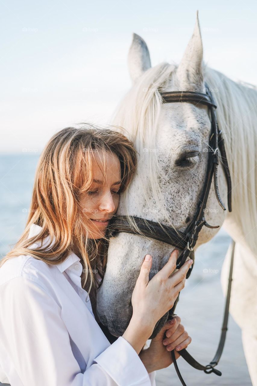 Young long hair woman in white shirt with white horse on seascape background