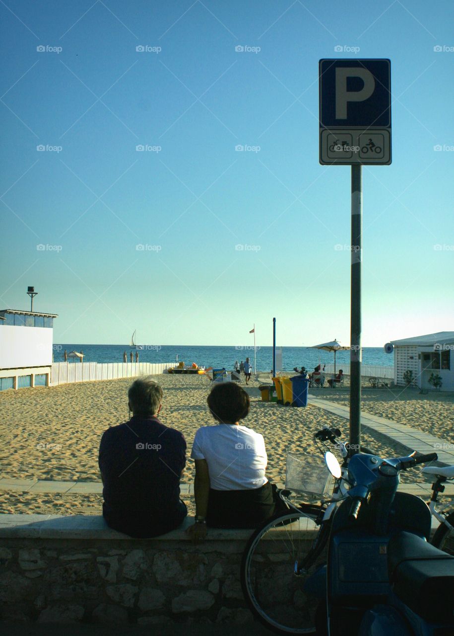 Middle-aged couple sitting on wall in front of beach, admiring the view and enjoying a warm Sunday.