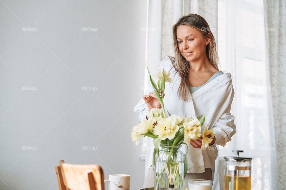 Young beautiful smiling woman forty year with blonde long hair in white shirt with bouquet of yellow flowers in hands near dinner table in bright interior at the home