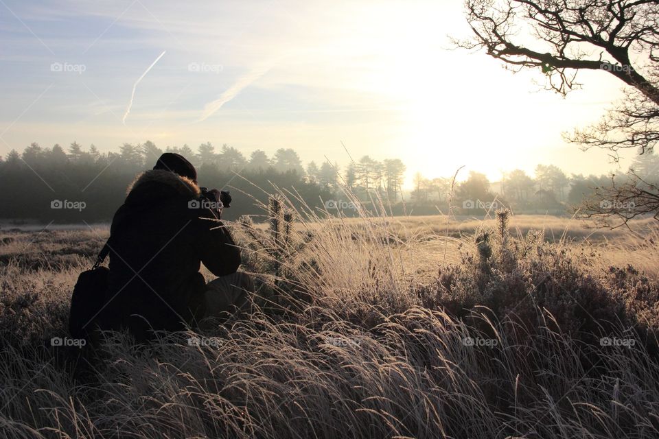 A portrait of a photographer sitting in the meadow taking pictures of the sunrise in winter.
