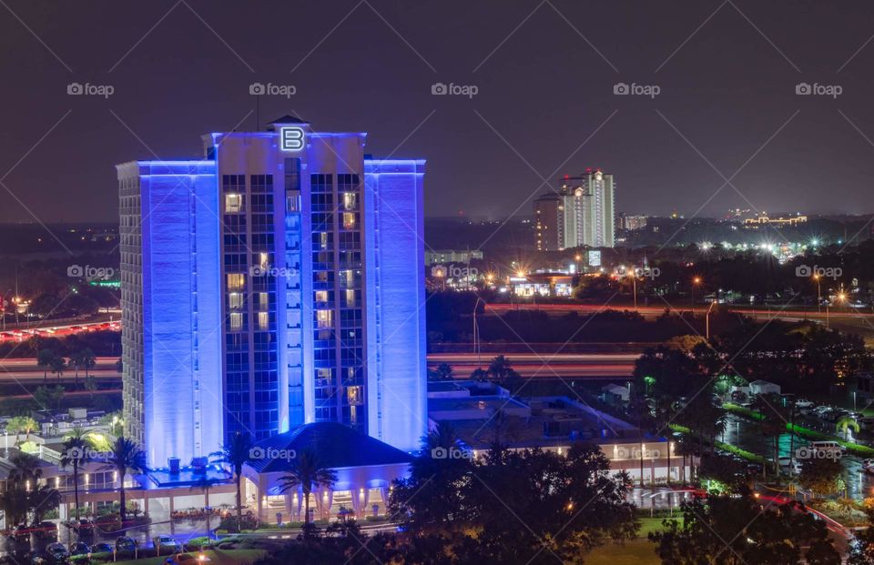 tall resort building colored bright blue by lights with surrounding area in foreground and city lights other tall resorts in background in a night sky