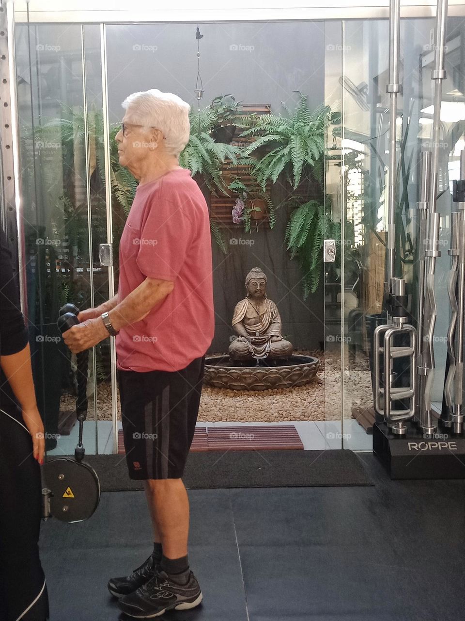 Elderly man at the gym, standing up working arms, gym with plants and a statue