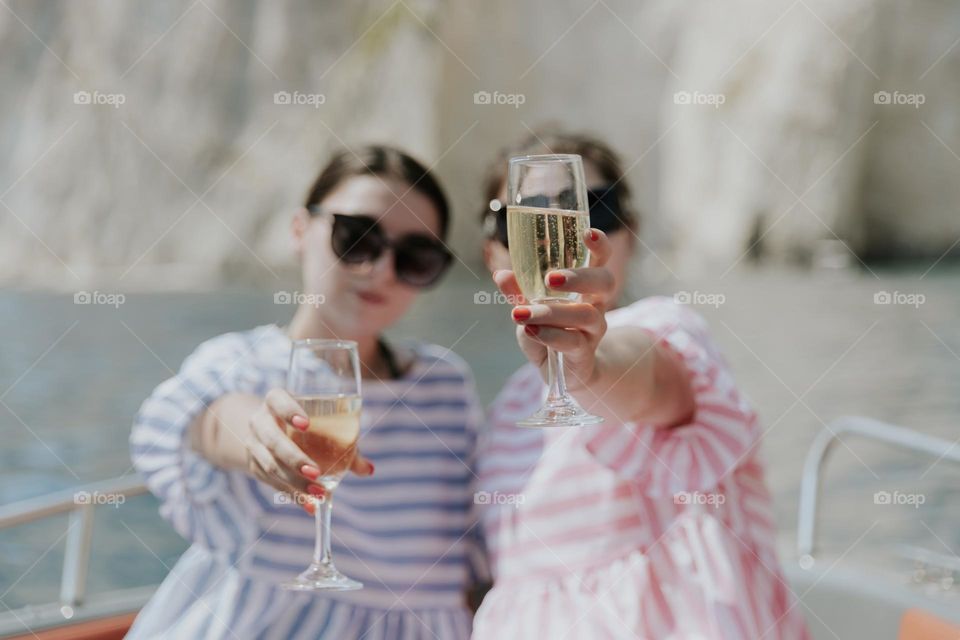 Two young and beautiful brunette girls in sunglasses hold glasses of champagne, holding them out, standing on a boat in the sea against the backdrop of blurred rocks, close-up side view.