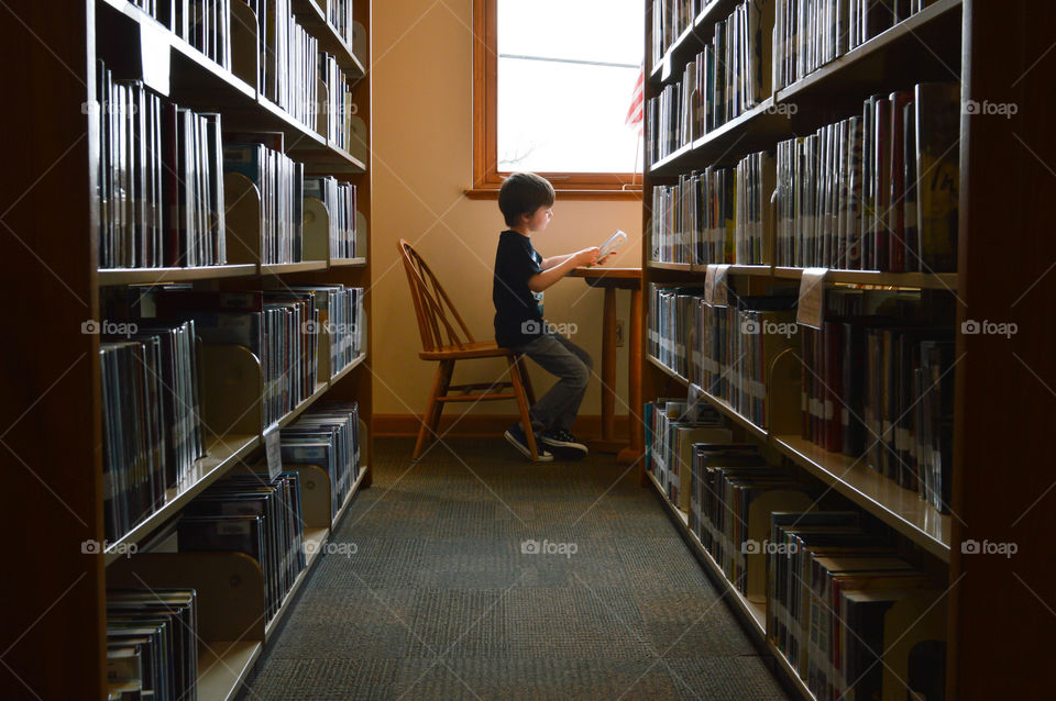 Two rows of books at a library with a young boy reading at a table at the end