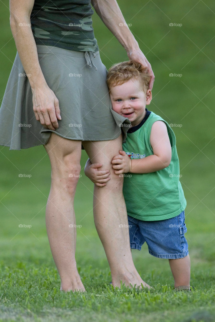 shy young boy holds on to his mothers leg at local park. shy young boy. red headed boy. by arizphotog