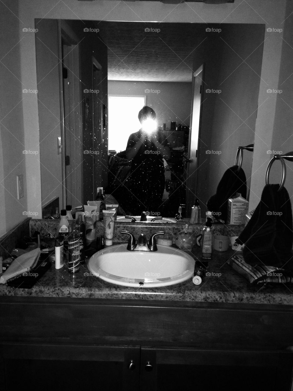 Short-haired person taking a mirror selfie at a cluttered bathroom sink. Flash obscures face, grayscale image.
