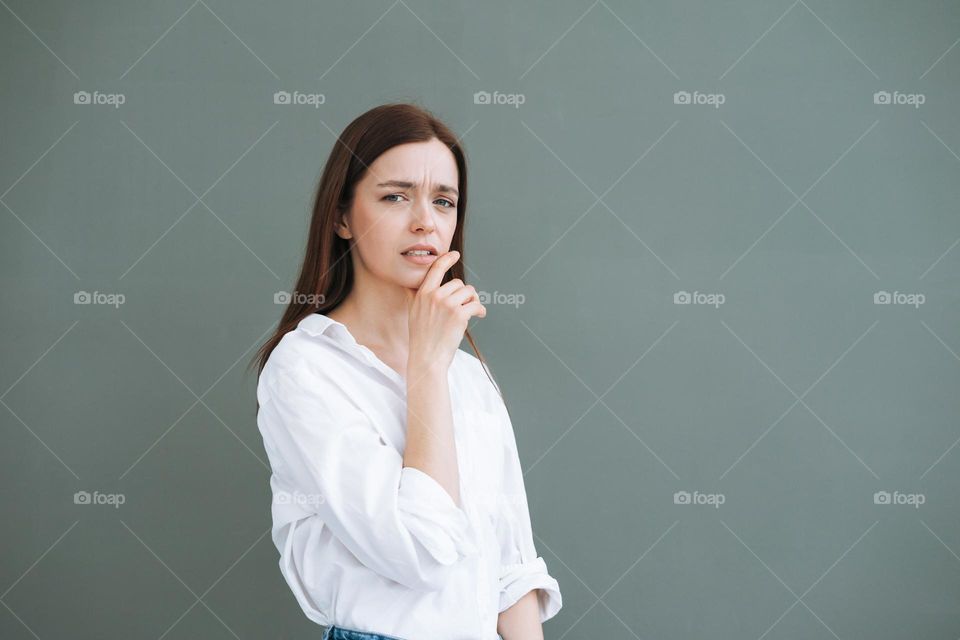 Young frown woman with long hair in white shirt on the grey background, negative emotion