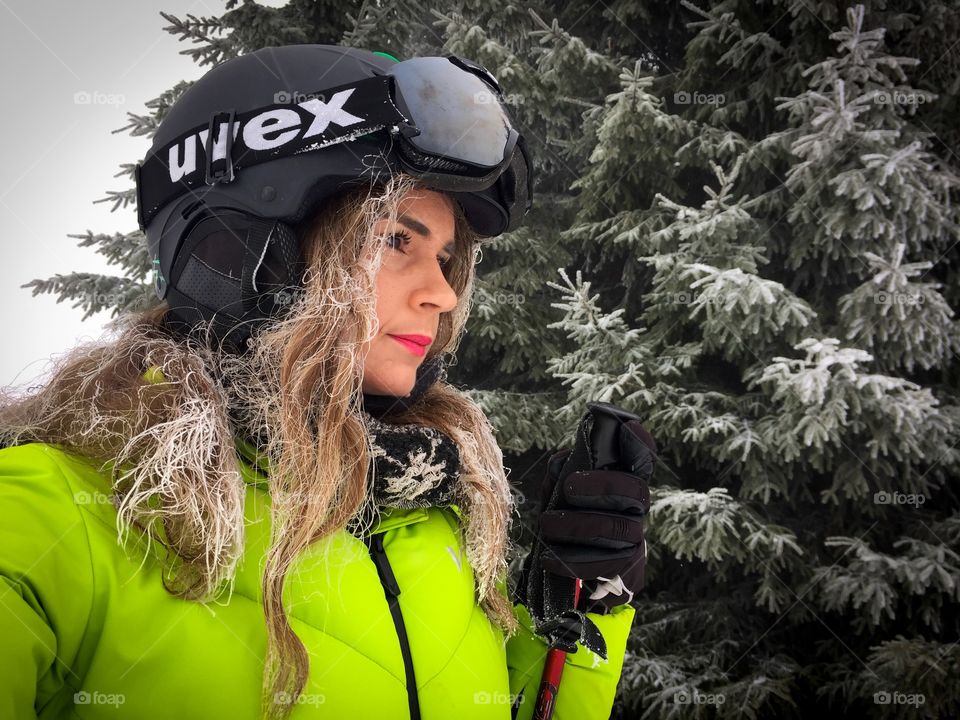 Portrait of woman wearing Uvex ski glasses and Uvex ski helmet and holding ski poles with pine cone trees covered in snow in the background