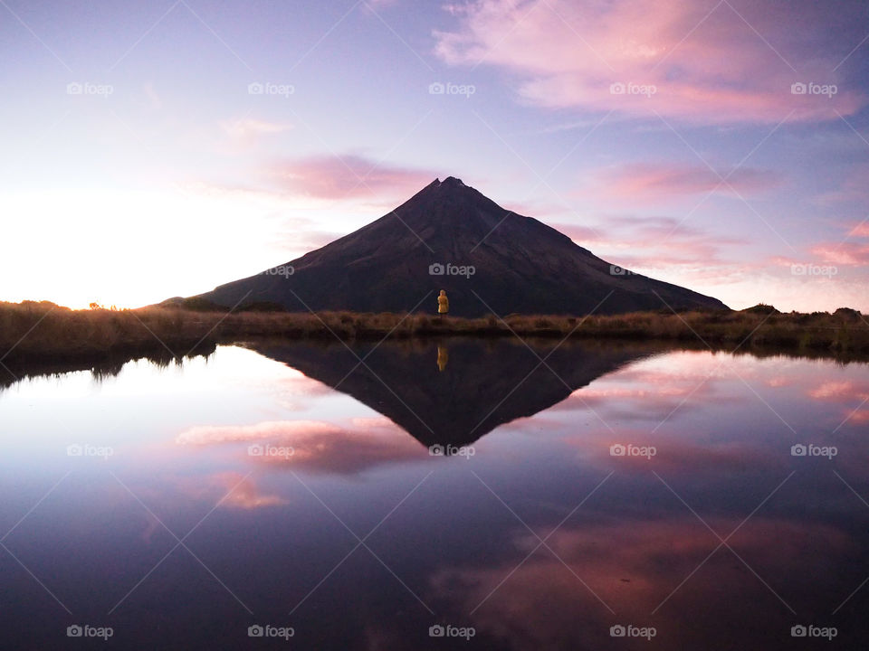 A single person stands in front of a mountain and is perfectly reflected on the lakes surface