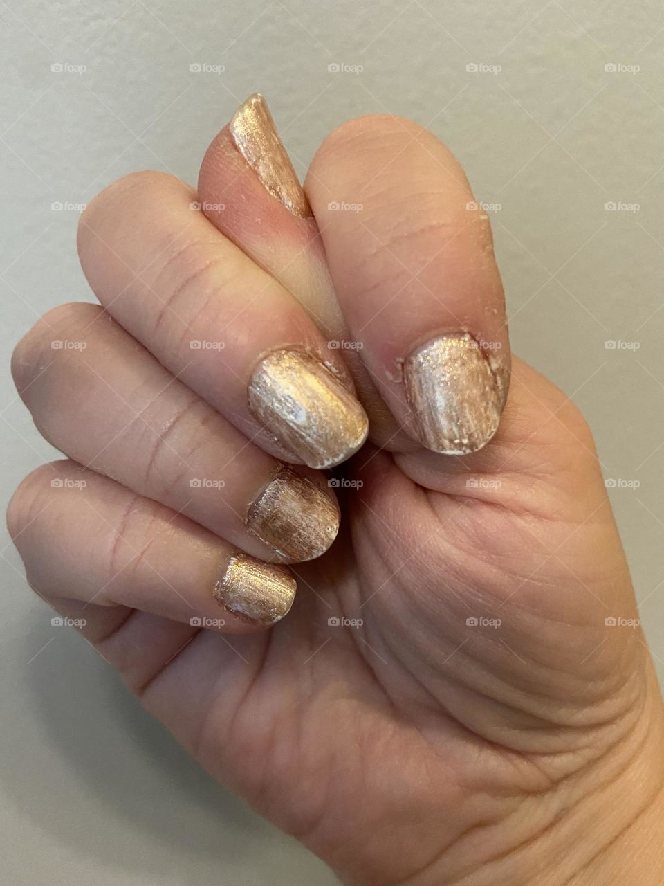 A hand in a fist with the thumb tucked in and peeking out, showing fingernails painted marbled-gold (a layer of white with a gold polish on top) against an off-white wall 