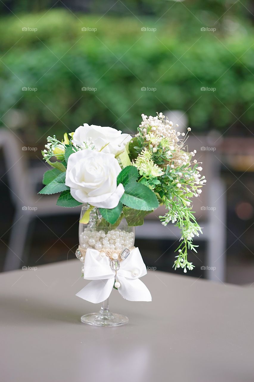 Wedding reception. A glass of white roses decoration at the wedding reception. White roses are artificial flowers, they made from fabric and plastic. Selective focus on the jewelry and the bow ribbon.