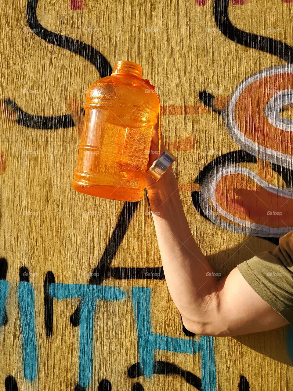 Arm holding orange water bottle against yellow graffiti art wall.