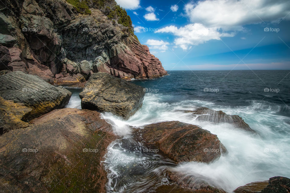 Waves crashing in on a rocky rugged coastline. Beautiful puffy clouds in a blue sky.