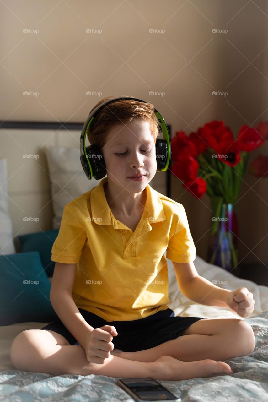 Close-up portrait of a child boy listening to music in headphones sits in the bedroom on the bed, music lover