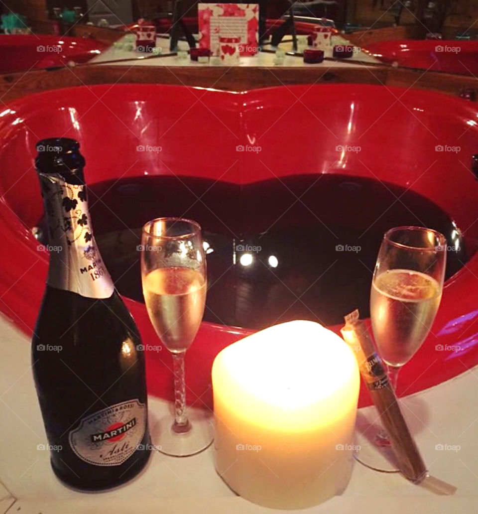 Red, filled heart shaped jacuzzi indoor hot tub with champagne, flutes, a lit candle, and a cigar, creating a romantic setting. 