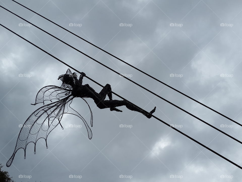Sculpted wire fairy climbing up power lines backed up by a dark grey stormy sky