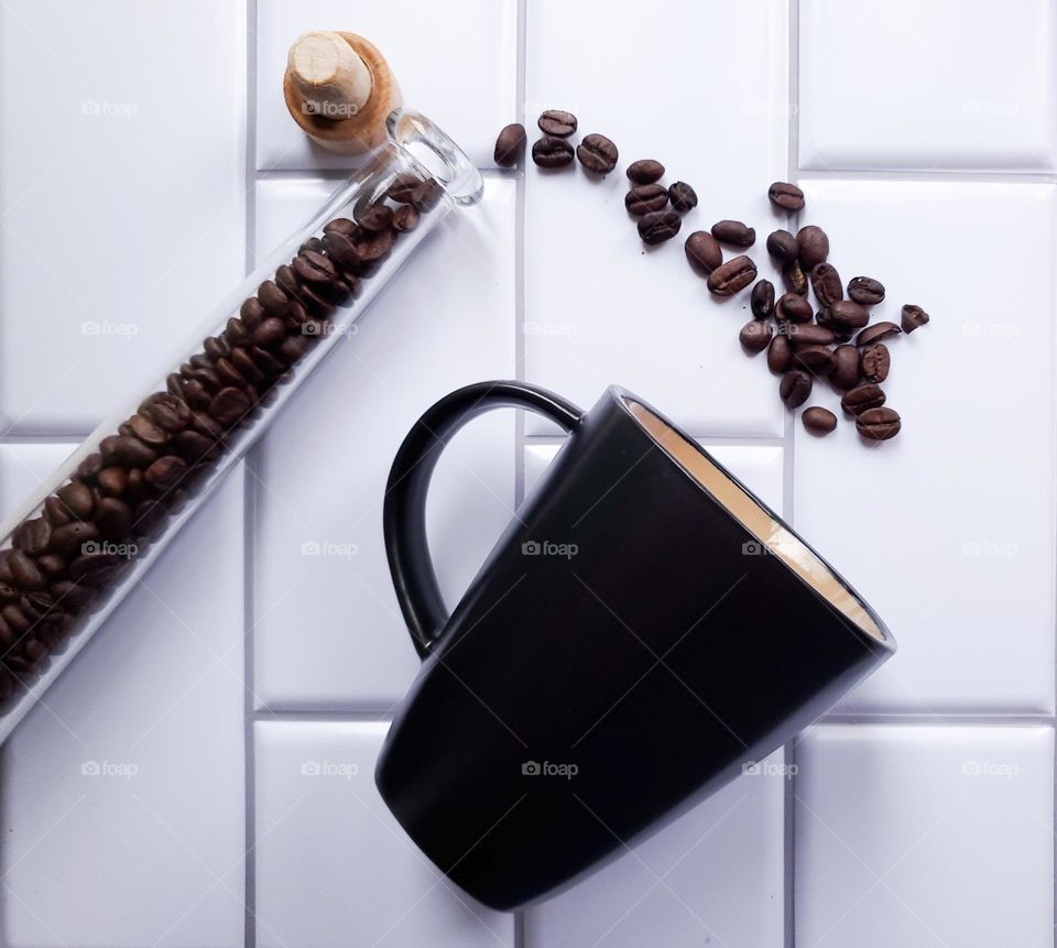 Coffee beans spilling out of slender glass container into a diagnolly tilted black coffee mug in front of a vertical white kitchen tile background.